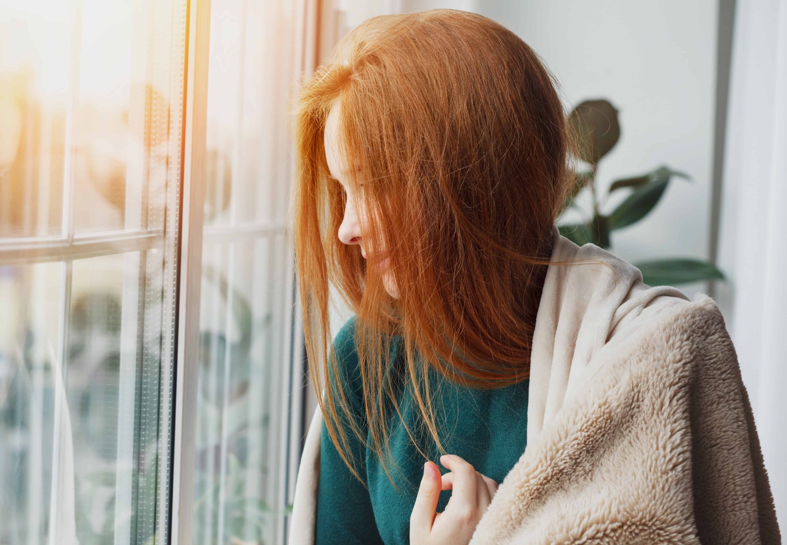 beautiful thoughtful pensive calm smiling young woman with long red hair looking through the window at home wrapped in warm comfy blanket